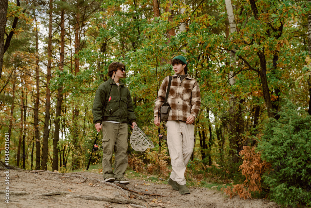 Naklejka premium Two friends walk through a vibrant autumn forest, preparing for a fishing trip. They carry fishing rods and a net as leaves fall around them, showcasing the beauty of nature.