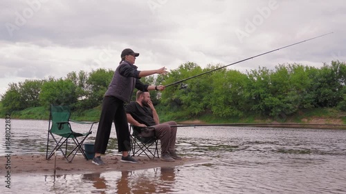 Woman standing on the riverbank casting a fishing line while a man sits beside her in a chair, both enjoying a relaxing day of fishing together outdoors in a beautiful natural setting
