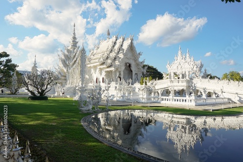 Majestic White Temple (Wat Rong Khun) in Chiang Rai, Thailand, featuring intricate architecture and a stunning mirror reflection on a calm pond under a clear blue sky.