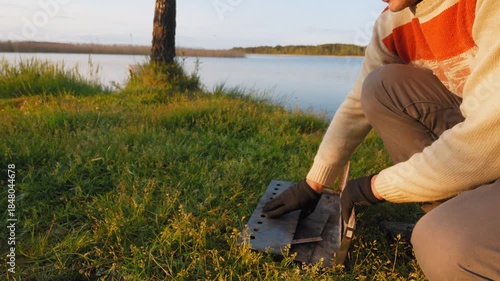 Young man assembling a portable barbecue grill on the green grass by the lake, preparing for an evening picnic and cooking delicious food in a beautiful natural landscape at sunset
