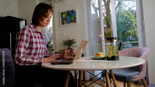 Smiling adult woman in works from home in sunny kitchen, typing on laptop, enjoying work-life balance, singing joyfully, feeling relaxed and inspired by remote job freedom.