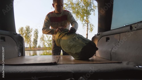 Young man unpacking and setting up a comfortable sleeping bag inside the trunk of his car, preparing to sleep while on a road trip adventure in a scenic lakeside location at sunset
