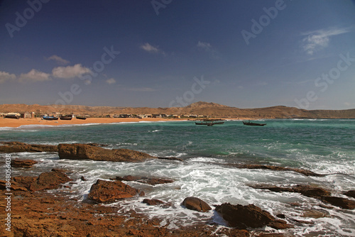 View of turquoise waves crashing on a rocky shore, contrasting with the sandy beach and distant structures under a clear sky, Karachi, Sindh, Pakistan.