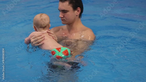 4K Smiling baby with toy in his hands swimming with swim coach in a swimming pool.