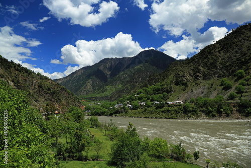 View of a rushing river snaking through a lush green valley beneath towering mountains under a bright blue sky with fluffy white clouds, Neelum Valley, Azad Kashmir, Pakistan.