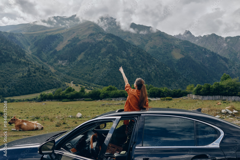 Fototapeta premium Woman in an orange jacket leans from a car window with arms raised, enjoying freedom in a vast mountainous landscape with a grazing cow and wide valley behind.