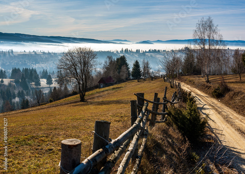 Countryside Mountain Road with Wooden Fence in Rural Landscape. Sunshine in the late autumn mountain landscape with a fence and dirt road above misty valleys, Harghita County, Livezi village, Romania.