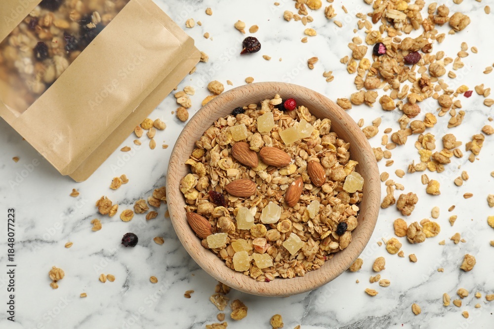 Naklejka premium Tasty granola with dried fruits and nuts in bowl and paper bag on white marble table, flat lay