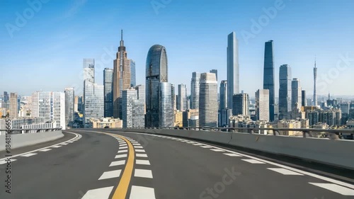 Empty modern highway leads toward a bustling urban skyline with diverse skyscrapers under a clear blue sky, showcasing urban architecture and cityscape.