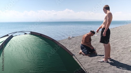 Two men set up a camping tent on the shore of the blue sea hammering pegs for guy lines holding the tent. Father and son spend a weekend together by the sea. Active family outdoor recreation