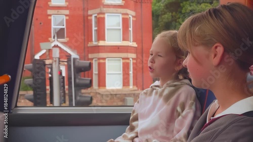 Two young friends travel on the top deck of a double-decker bus. They watch the urban English landscape unfold through the window, sharing a quiet moment on their city journey