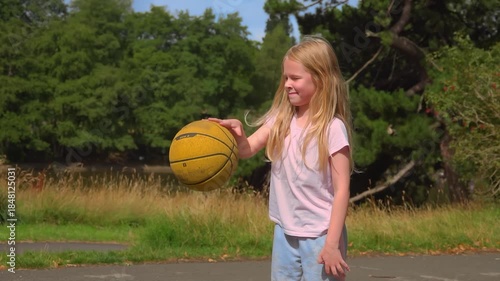 A little girl is making her first steps in learning basketball. She is uncertainly and hesitantly bouncing the ball on the ground, trying to master a new skill