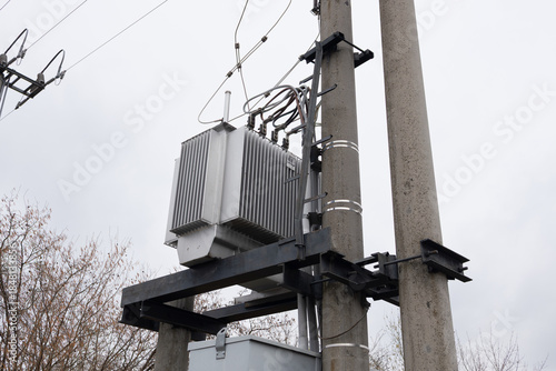Electrical transformer mounted on utility pole with wires and cables connected, surrounded by bare trees and cloudy sky in urban area during daytime