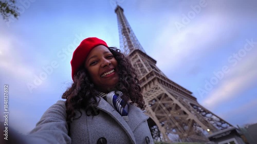 African millennial smiles at the camera while taking a selfie beside the Eiffel Tower in Paris at blue hour, wearing a red beret and coat, capturing urban travel vibes with direct gaze.