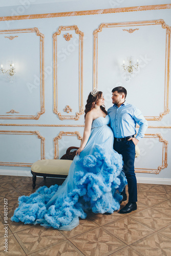 a married couple stands together near a beige sofa. married couple looking at each other. pregnant girl in a lush blue dress. young man in a blue shirt. family photo session. 