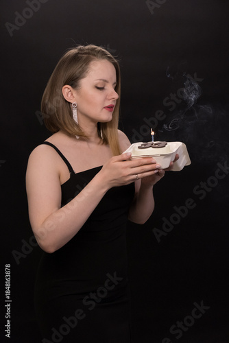A woman in a black dress blows out a candle on a bento cake. Smoke on a black background. A girl's birthday, happiness, a gift. High quality photo