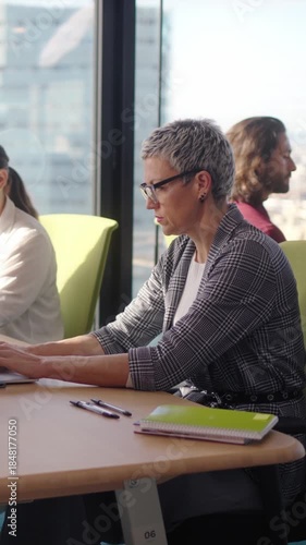Vertical. Middle aged professional works at an open plan desk in a bright corporate office, concentrating on tasks at a dual monitor setup with paperwork and notes by the window overlooking the city.