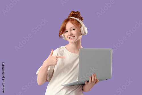 Cute schoolgirl in headphones pointing at modern laptop on lilac background