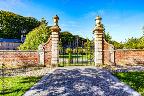 Cobblestone path towards gate with wrought iron gate between two stone pillars en brick walls, farm of Averbode Abbey, leafy trees in background, sunny day in Scherpenheuvel-Zichem, Belgium