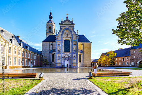 Exterior esplanade with impressive facade of Averbode Abbey, pond with reflection on water surface, standing woman, Baroque style, sculptures and bell tower, sunny autumn day in Belgium