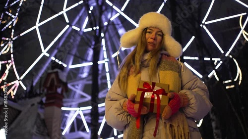 Cheerful young woman holding a present with a red ribbon while standing in front of a brightly lit ferris wheel at night during a snowy winter evening in a festive amusement park