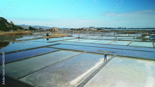 Drone view of geometric solar evaporation ponds with Asian worker collecting sea salt. Traditional salt farm landscape with reflections in shallow water, coastal Vietnam.