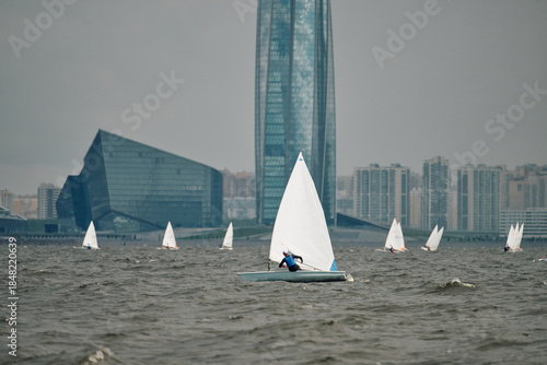 The sports sailing dinghy is heeling at cloudy day, athletes tilt the boat, speed and spray, reflection of sun on water surface, Russia, St. Petersburg