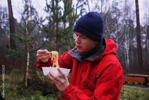 A young man eating instant noodles in nature. Lunch during a hike in the forest, fast food at a picnic