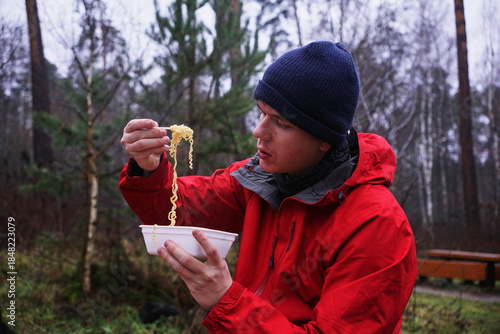 A young man eating instant noodles in nature. Lunch during a hike in the forest, fast food at a picnic