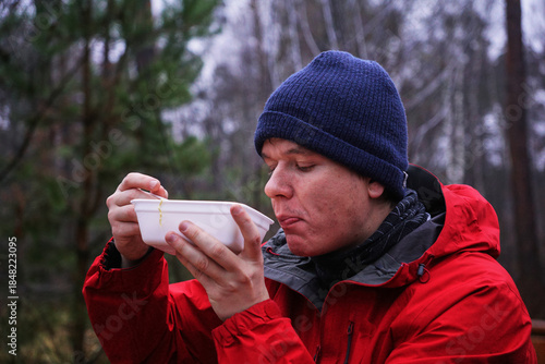 A young man eating instant noodles in nature. Lunch during a hike in the forest, fast food at a picnic
