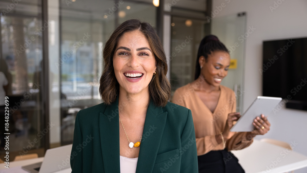 Obraz premium Happy beautiful Hispanic businesswoman posing for head shot in office workspace with diverse colleagues working behind, looking at camera, smiling. Banner portrait of confident female business leader