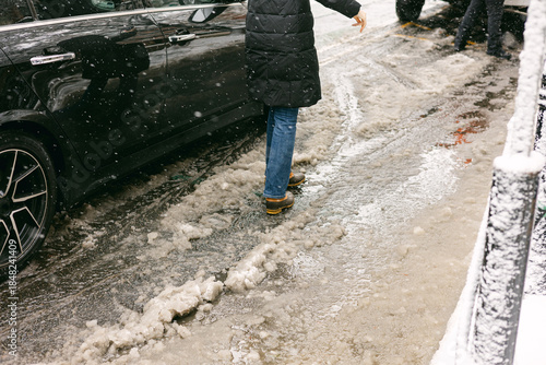 Person walks on icy street beside parked car during winter snowfall in urban setting