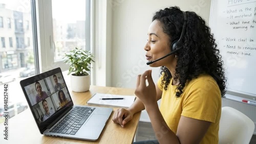 A focused and engaging young woman with curly hair actively participates in a virtual video conference from a bright, modern home office setting. She wears a professional headset with a microphone, ge