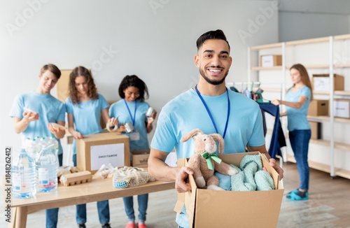 Fotografia In a charity center, an Arab male volunteer holds a box filled with clothes and toys while smiling