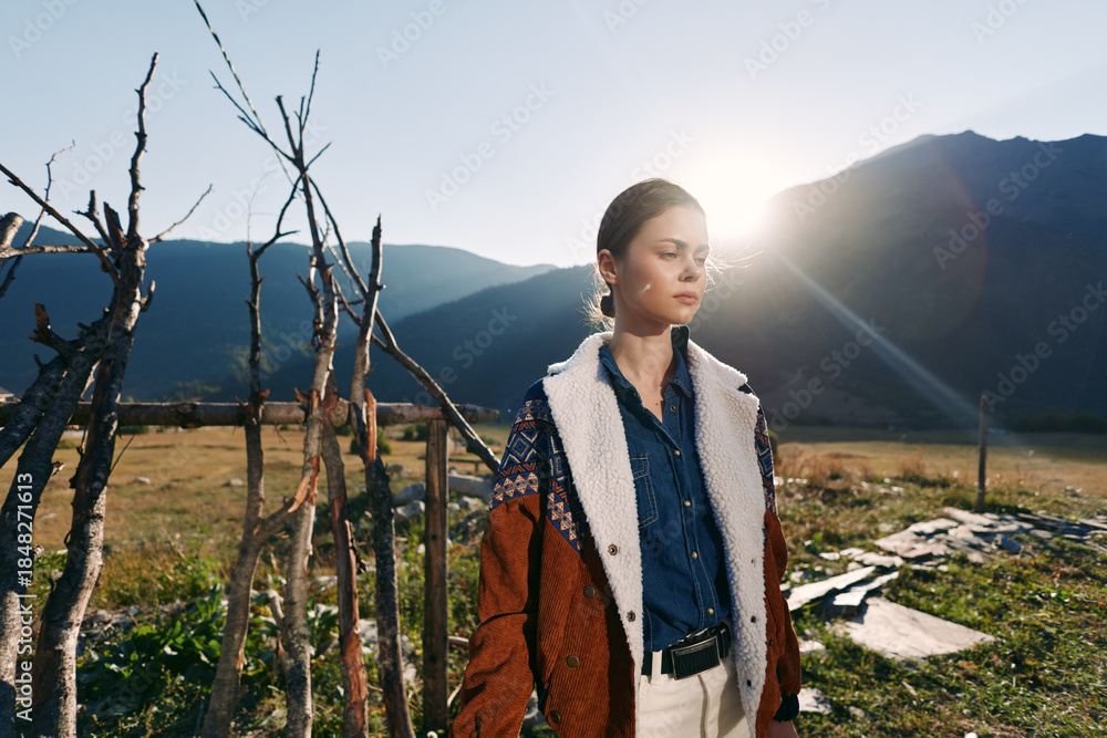 Naklejka premium Woman portrait in mountains with warm sunlight, fashion outfit and layered jacket standing outdoors by rustic wooden fence, serene landscape and travel lifestyle mood.