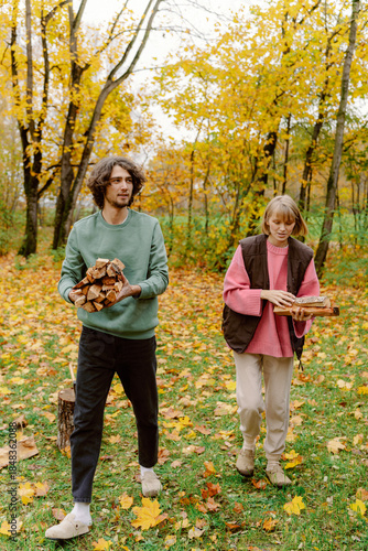 In a vibrant autumn forest, a couple collects firewood. The ground is covered in colorful fallen leaves, and trees create a warm backdrop. Their cozy attire adds to the seasonal charm.