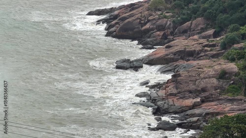 Rough surf crashes along uneven coastal rocks forming swirling white foam as faint patches of natural green higher on the shore add balance to the otherwise dramatic storm filled seascape. Wild
