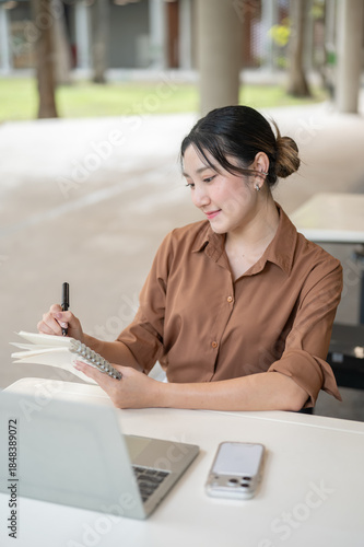 Asian woman student freelancer writing in notebook over laptop sitting at table in open air building
