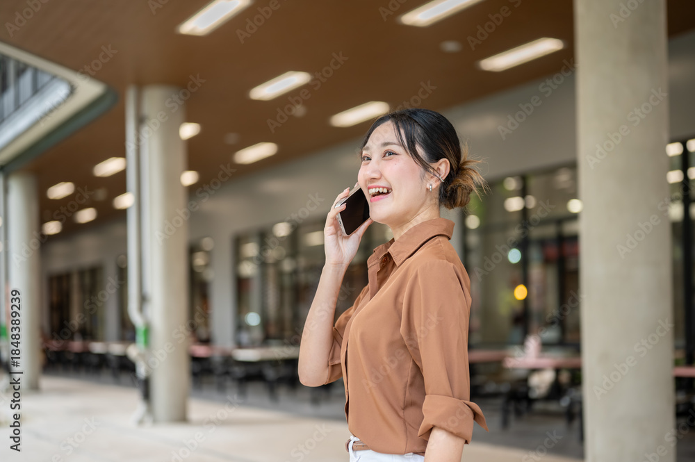 Fototapeta premium Asian woman student talking on smartphone looking ahead while standing sideway in open air building.