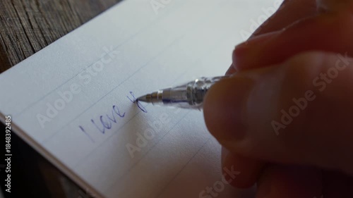 A macro style view reveals a woman writing the words I love you in a notebook with a blue pen, each slow stroke underscoring emotional honesty, personal meaning, and gentle connection. Written