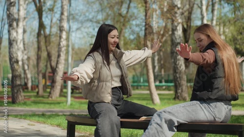 two women share smiles on park bench, asian and caucasian women revel in spontaneous outdoor joy, female friends from different backgrounds enjoy cheerful moments in open air together