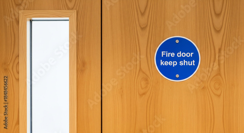 A wooden fire door with a window and a blue sign that says fire door keep shut on the right side