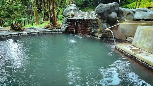A tranquil outdoor pool with turquoise water surrounded by large rocks and lush greenery in a natural setting with water flowing out of pipes into the pool.