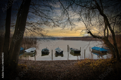 A peaceful lakeside scene at sunset with several small boats moored along a wooden dock, framed by bare tree branches and autumn foliage.
