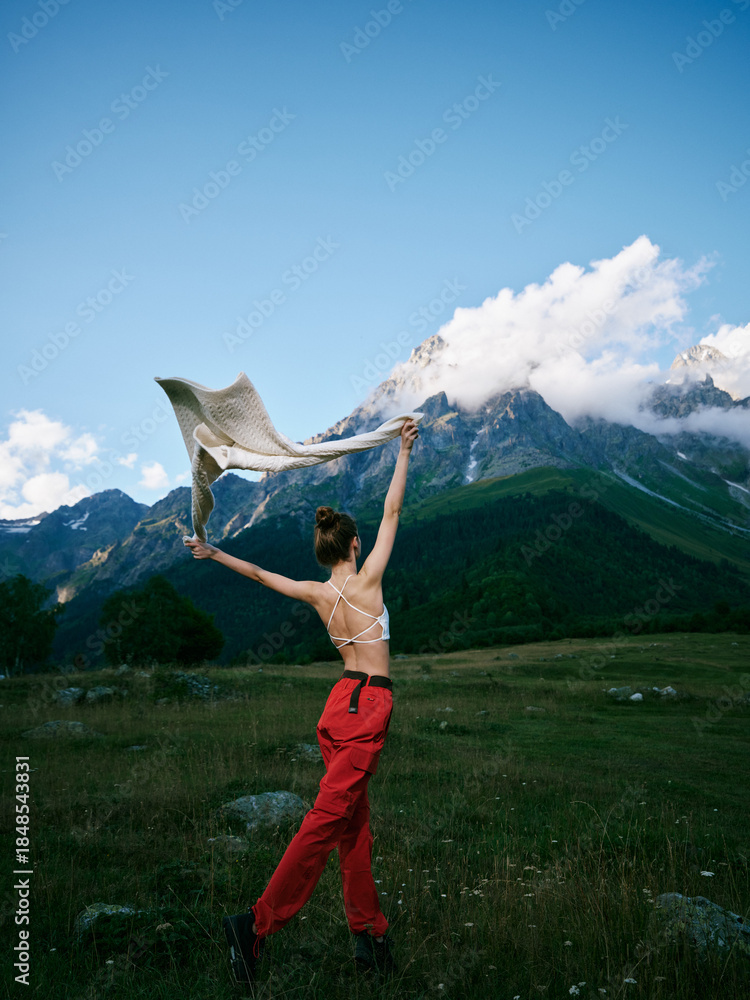 Fototapeta premium Dancer leaping in red pants with scarf in an open meadow beneath towering mountains, wind catching fabric, showcasing movement, freedom, and outdoor adventure