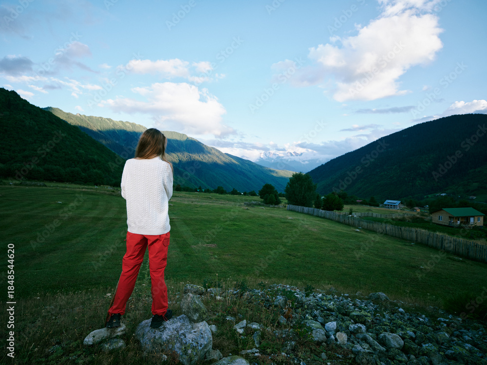 Fototapeta premium Woman standing on rocks overlooking a green valley with mountains in the distance, clear blue sky and scattered clouds, capturing a serene outdoor landscape scene with calm rural vibes.