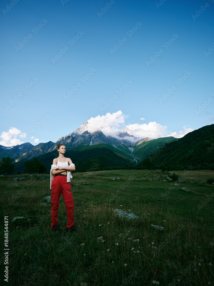 Naklejka premium Woman stands in a mountain meadow with bold redpants, under a clear blue sky, capturing outdoor adventure and serene nature amid alpine scenery