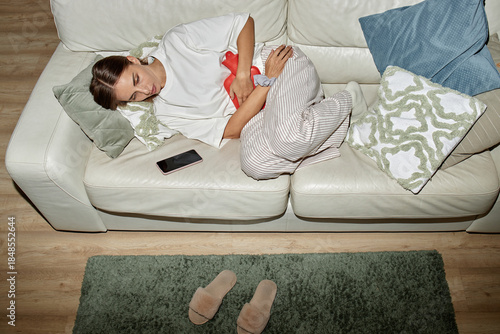 Bild auf Leinwand Caucasian young adult woman lying on sofa clutching stomach with pained expressi