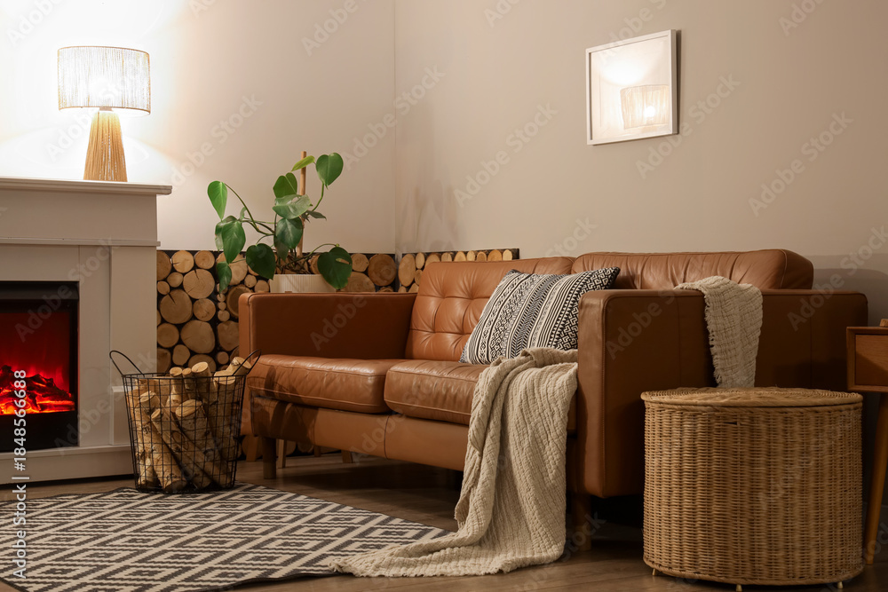 Fototapeta premium Interior of living room with sofa, glowing lamp, wicker table and firewood near fireplace in evening