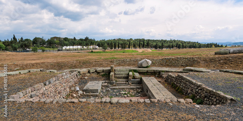 Pergamon Asclepieion ruins, Bergama, Izmir, Turkey. Ancient stone structures, dry fields, and trees under a cloudy sky. Historical archaeological site.